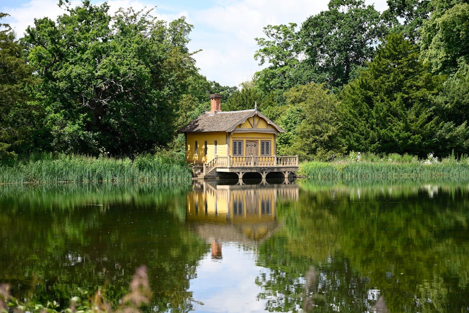 A small yellow wooden house with a thatched roof and decorative window panes, situated on the edge of a calm lake surrounded by dense green trees and tall grass, reflecting clearly on the water's surface. The house is accessible via a short wooden deck with a railing, and the background features a lush, wooded landscape under a partly cloudy sky. This image captures a tranquil setting that could be part of a house move or relocation process, with no visible furniture or moving equipment present, emphasizing the natural environment typically associated with property locations in the Watford area, as seen in the Cassiobury Park removals guide at watfordmanandvan.com, a company specializing in removals and home relocation services.