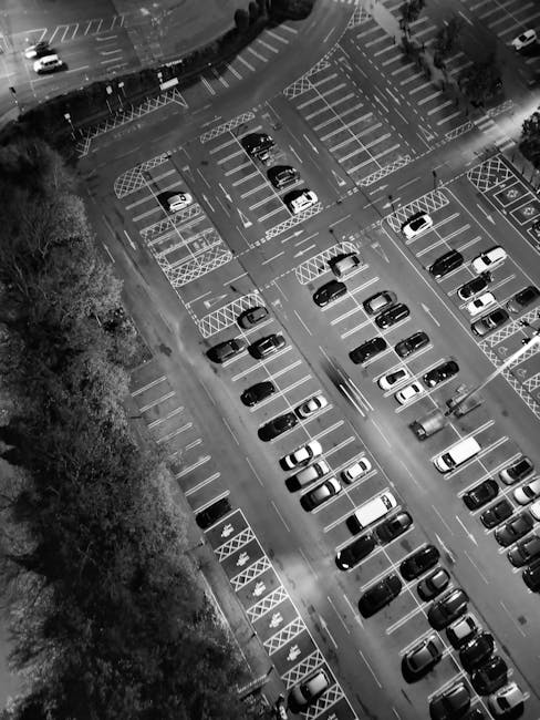 Aerial view of a large multi-storey parking lot with numerous parked cars arranged in rows, including small hatchbacks, sedans, and SUVs, with some spaces marked for disabled parking with blue signage. The parking area is bordered by trees along one side, and the lot features clearly delineated parking bays, some of which are empty. The surface of the lot appears weathered, with visible painted lines and markings. The image is in black and white, emphasizing the organized layout of vehicles and space allocation, and it reflects a typical urban parking environment suitable for residential or commercial building proximity, relevant to house removals and transport logistics, as might be discussed at Watford Man and Van during home relocation projects such as those described in the Cassiobury Park removals guide.