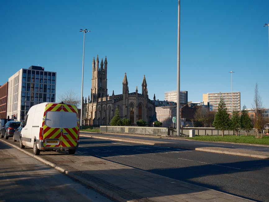 Two men are engaged in a home relocation process outdoors, near a brick wall and a paved driveway, with a white delivery van parked beside them. They are loading a large cardboard box into the back of