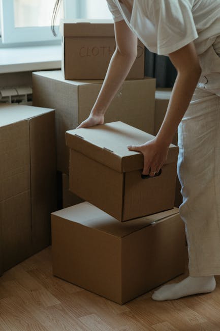 A person wearing light-colored clothing is in an indoor space, carefully placing a medium-sized cardboard box onto a stack of similar boxes during a home relocation process. Several other cardboard bo